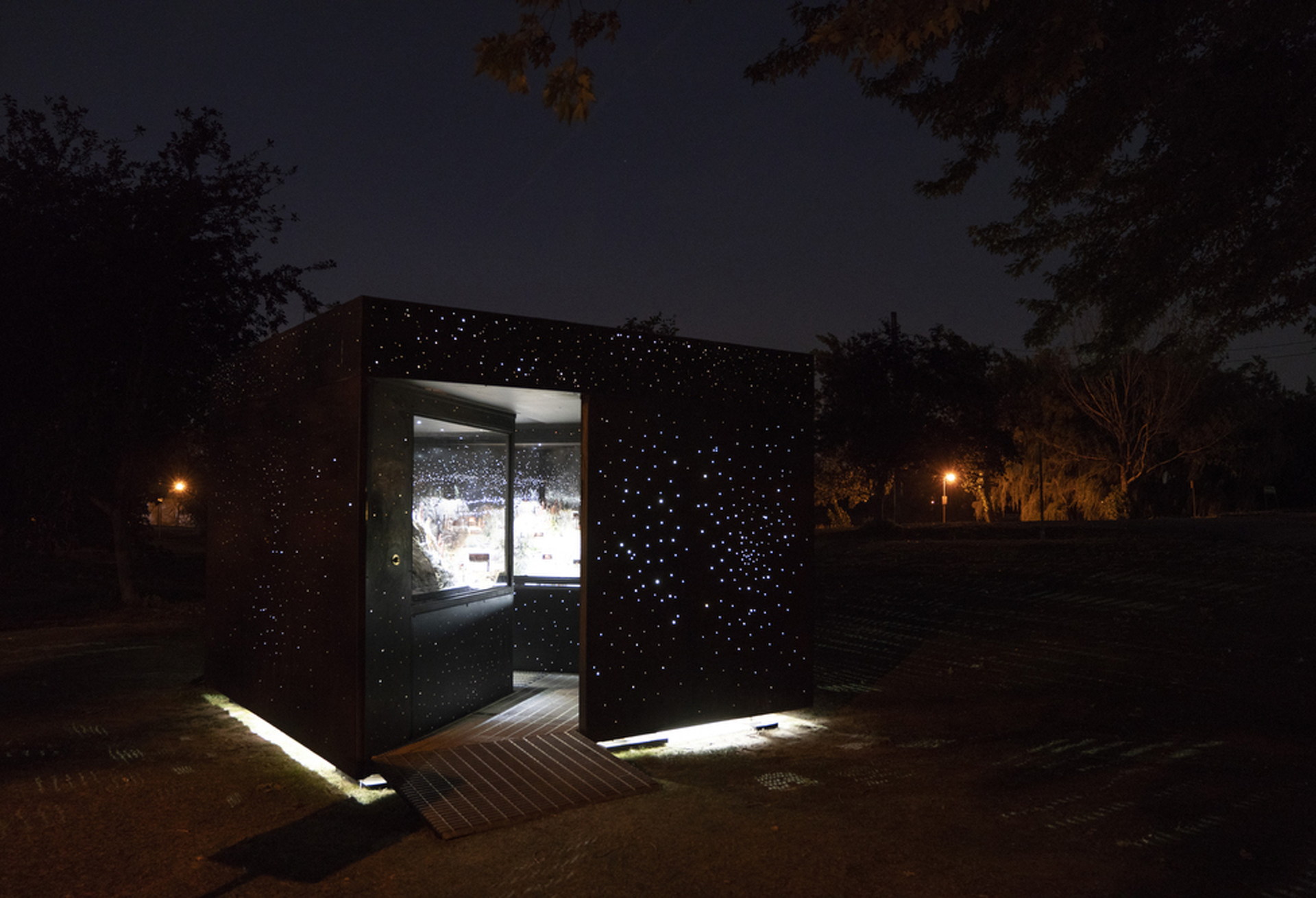 Set in a park, a path leads into a dark box with glass cases and small holes emitting light.