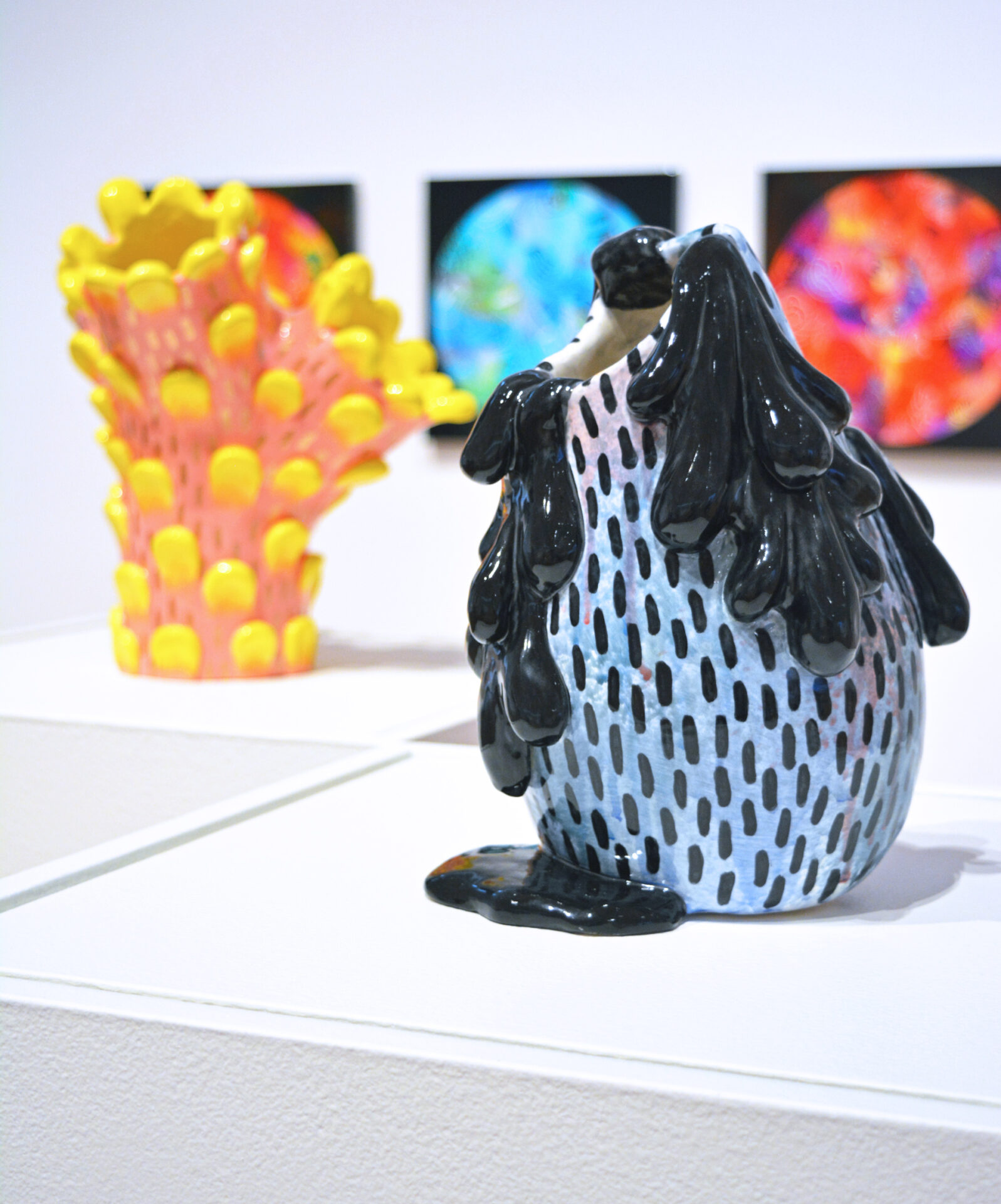 Two brightly coloured ceramic vessels. The vessel in the foreground is blue and black, resembling a jug with black water drips. The vessel in the background is pink and yellow and resembles coral or an abstract floral form.