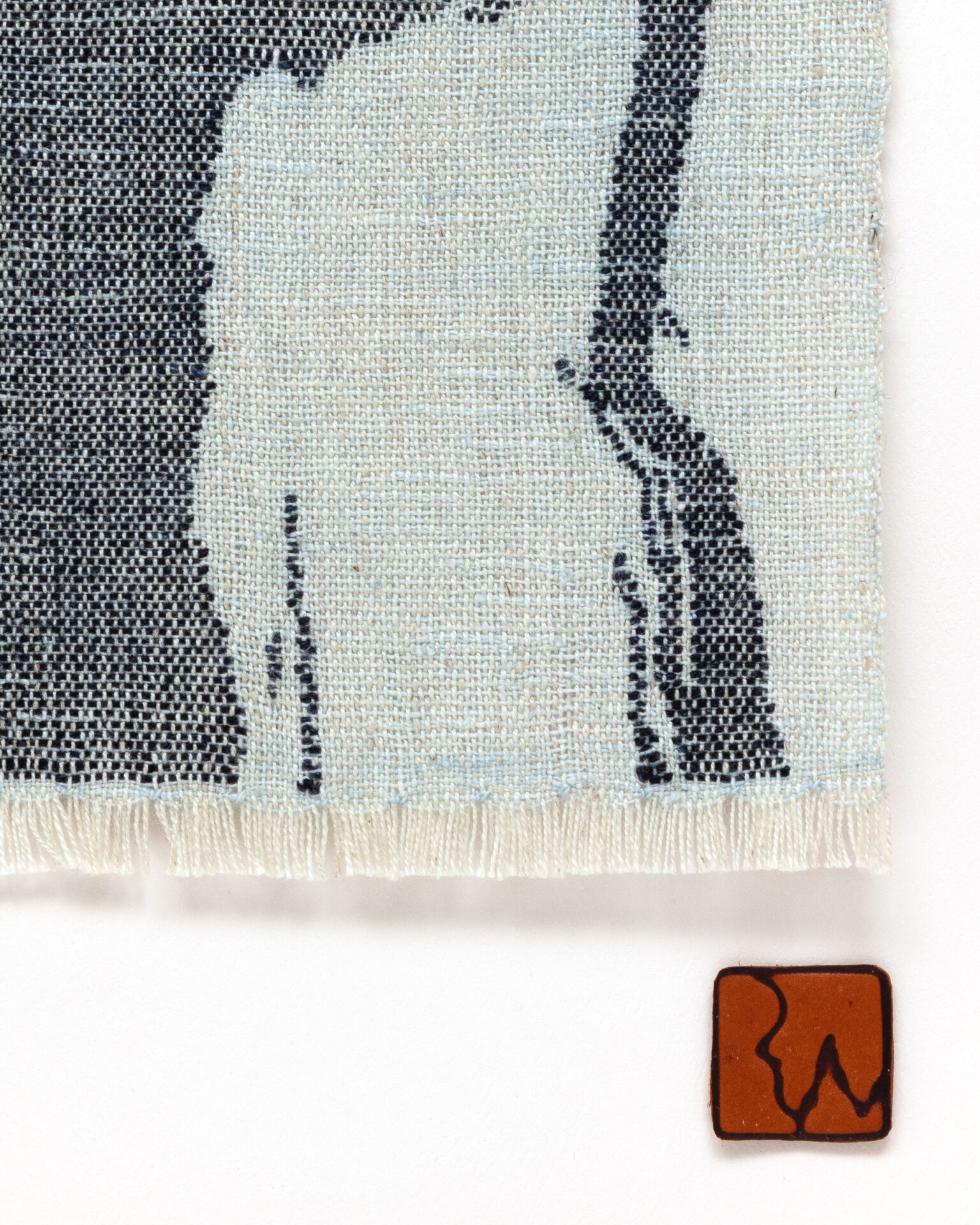 Detail of abstract woven fabric art showing navy blue shapes in a pale blue cloth, small fringe and leather patch for the artist’s signature.