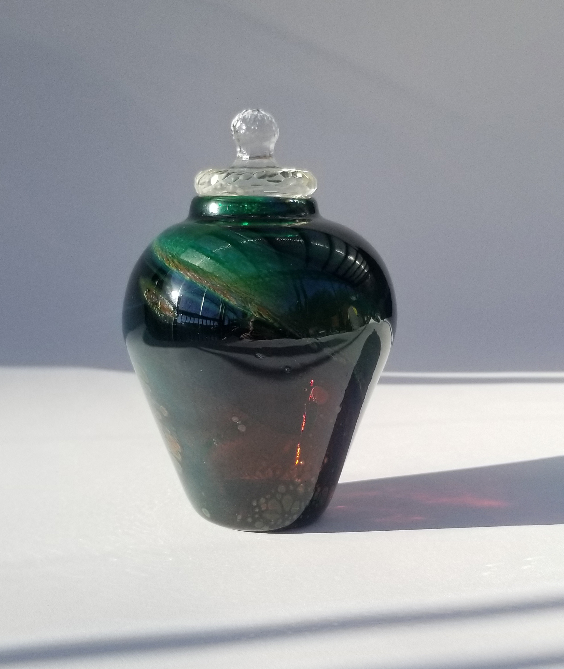 Image of a glass jar with a wide shoulder and a tapered bottom against a light grey background. The jar has a clear cut crystal lid that sits atop a small neck. The jar is dark green and black with flecks of ruby red.