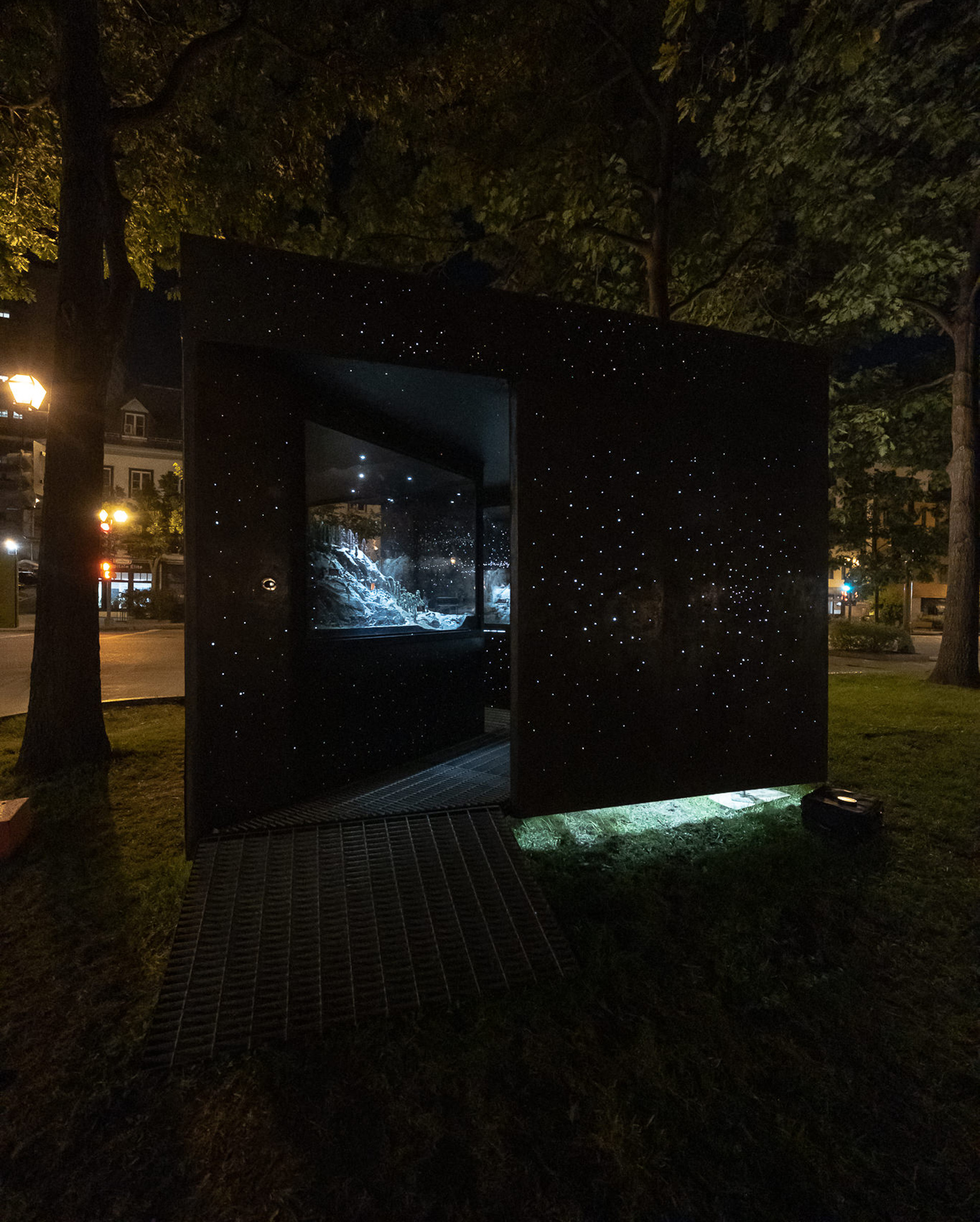 Set in a park, a path leads into a dark box with glass cases and small holes emitting light.