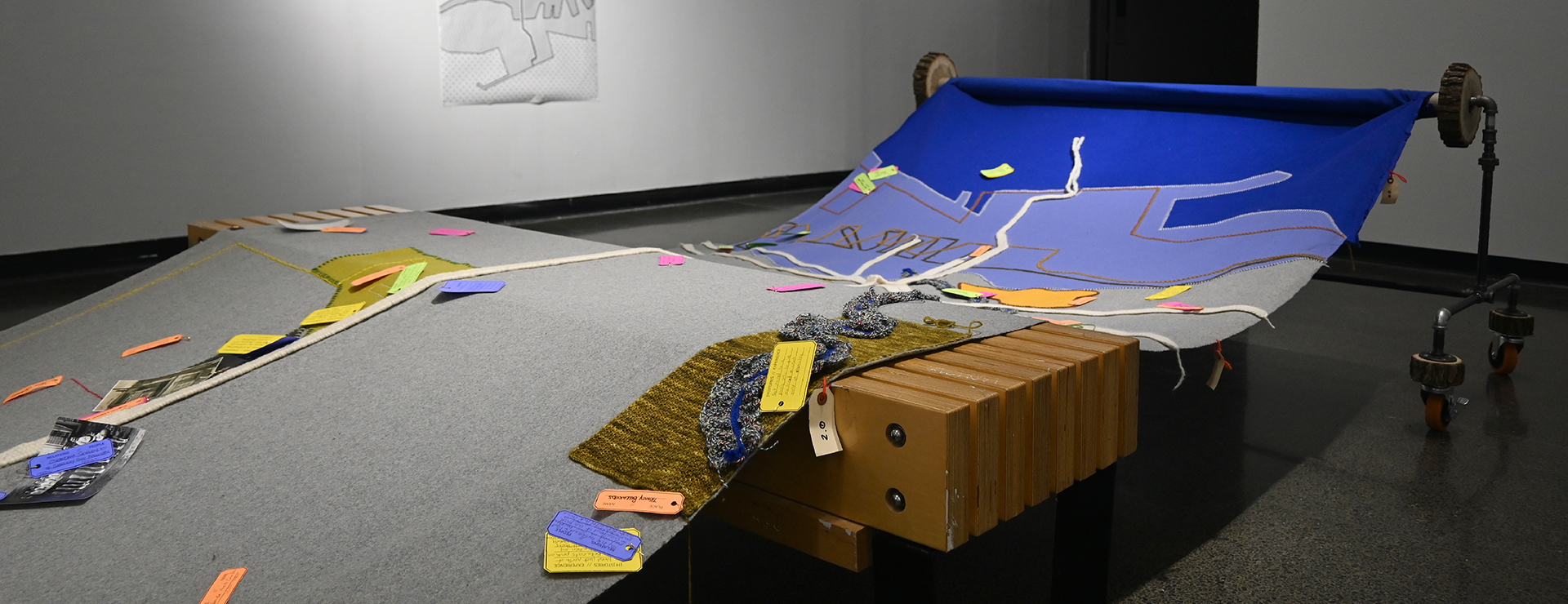 Large textile map made of grey and blue fabric, with multi-coloured tags. The Map is supported by a steel pipe structure and is installed in a gallery space.