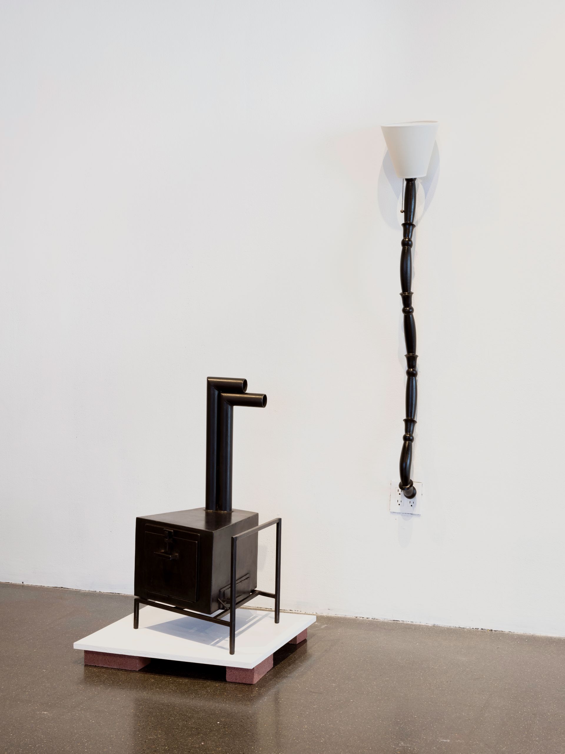 A black, geometric stove and a black, sconce that plugs into the wall and looks. like a vine against a white gallery wall.