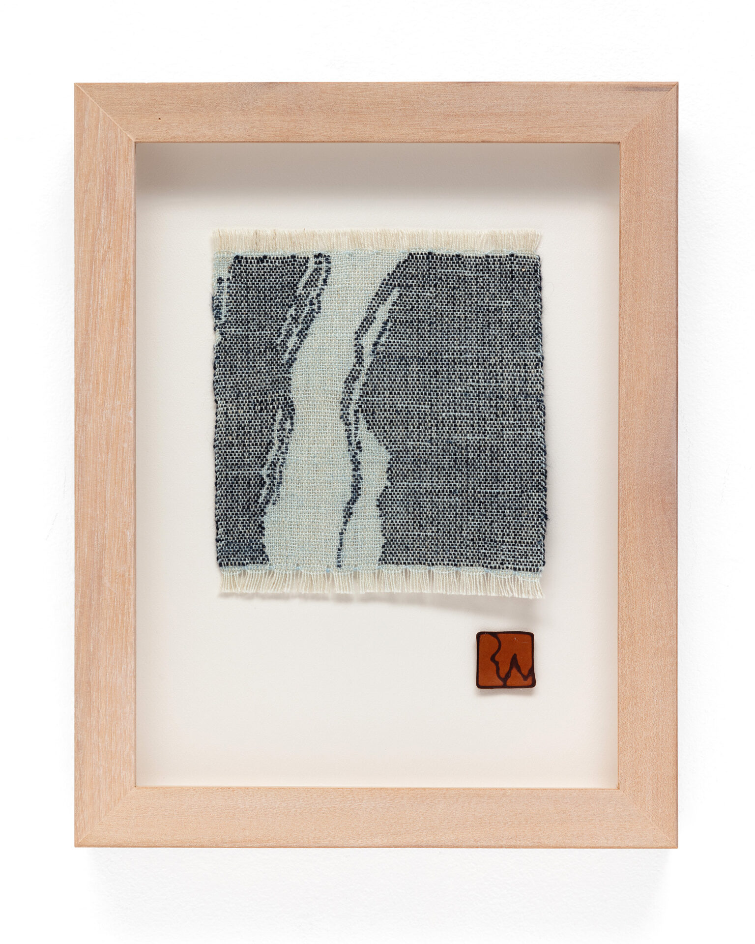 Piece of abstract woven fabric art showing navy blue shapes in a pale blue cloth. The weaving is mounted in a pale wooden frame with a brown leather patch for the artist’s signature at bottom right.