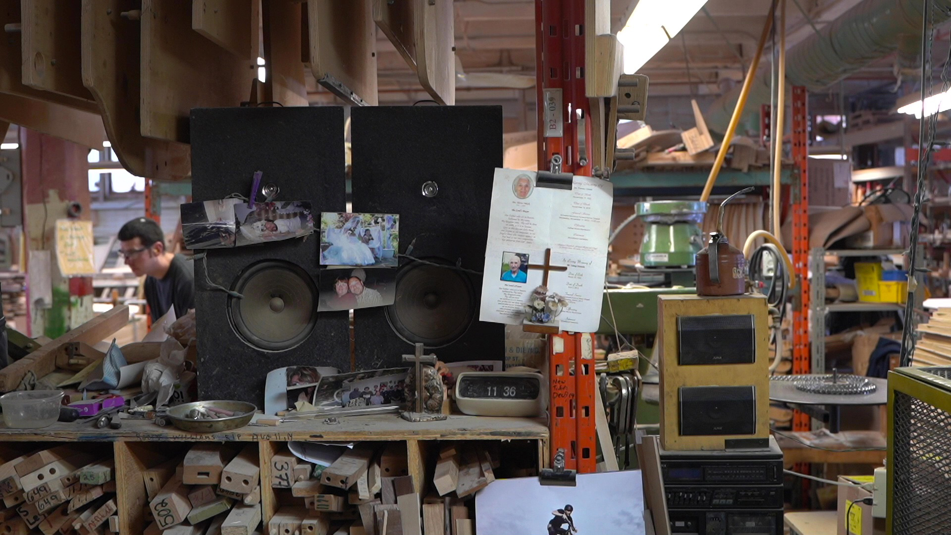 Factory scene with music speakers, photos of workers families pinned up, wood organized, worker in the background