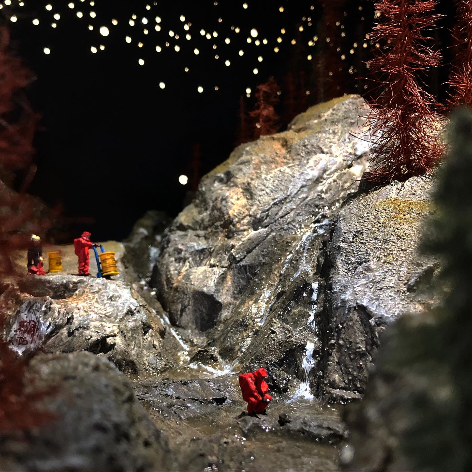 Diorama of rocky terrain with people in red outfits moving yellow barrels.