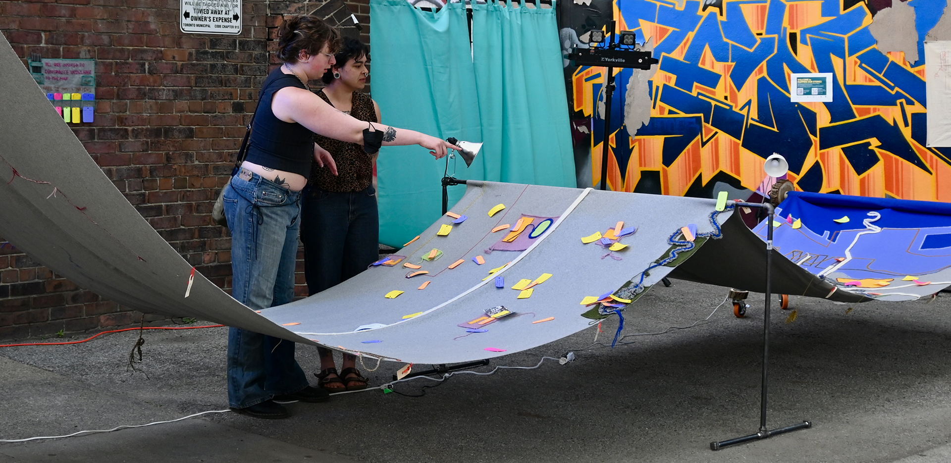 Two people interacting with a large textile map made out of grey and blue fabric, with multi-coloured tags. The Map is supported by a steel pipe structure and is installed next to a brick building and brightly coloured mural.