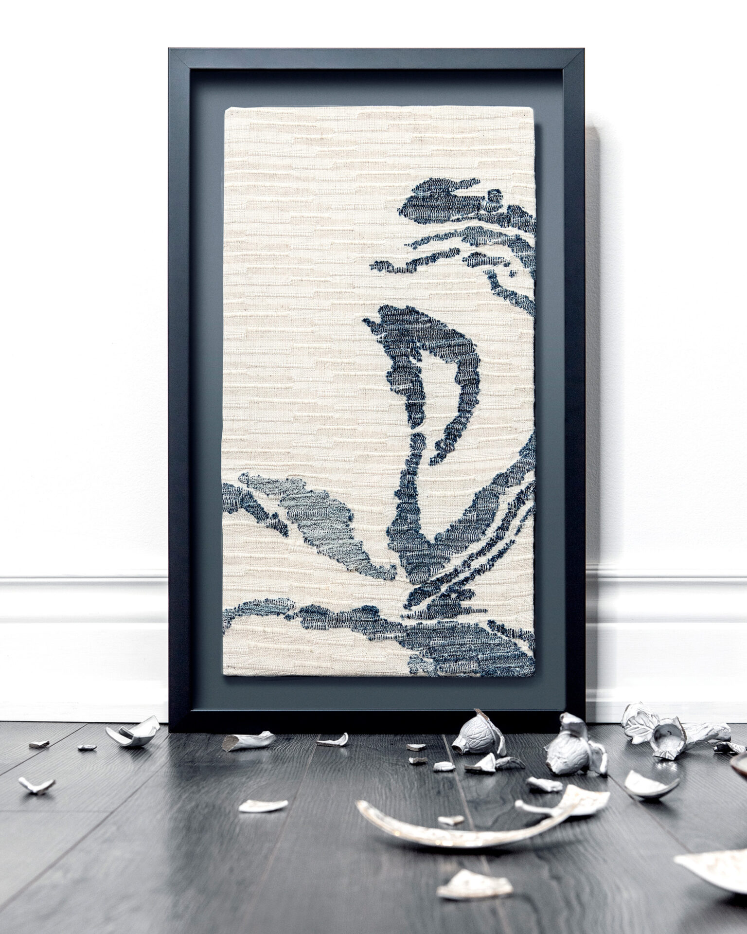 Tall piece of abstract woven fabric art showing navy blue shapes that swirl on a beige background. The frame is surrounded by shards of broken ceramics.