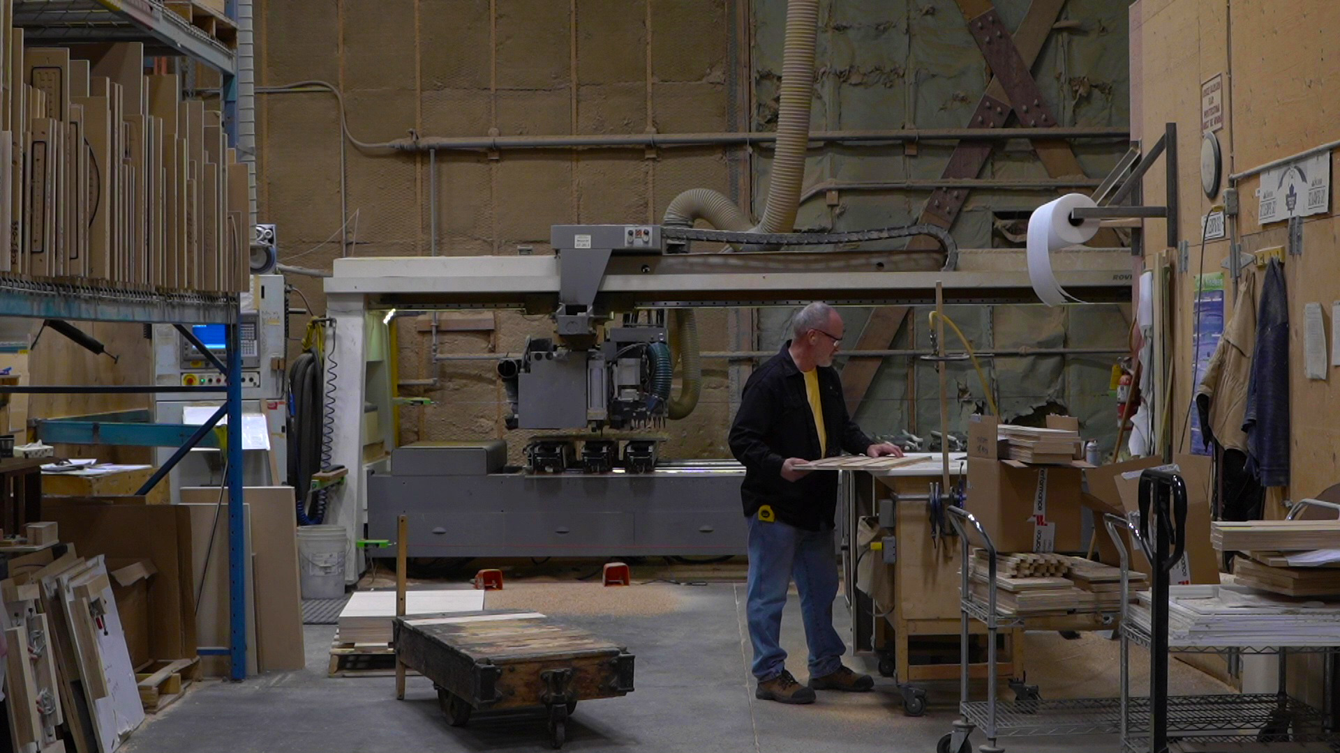 Factory scene of open workspace with worker in the centre, organized wood for making chairs, big industrial saw in background